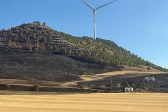 Una tierra de labranza y la ladera de un cerro calcinadas junto a la entrada de varias bodegas con un aerogenerador al fondo.