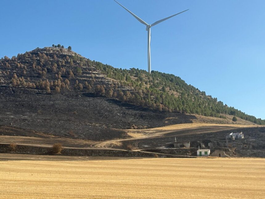 Una tierra de labranza y la ladera de un cerro calcinadas junto a la entrada de varias bodegas con un aerogenerador al fondo.