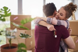 Una pareja se abraza mientras ella sostiene en las manos las llaves de una casa y al fondo se ven cajas de mudanza