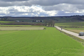 Furgoneta circulando por una carretera provincial entre campos de cultivos con un conjunto de casa al fondo.