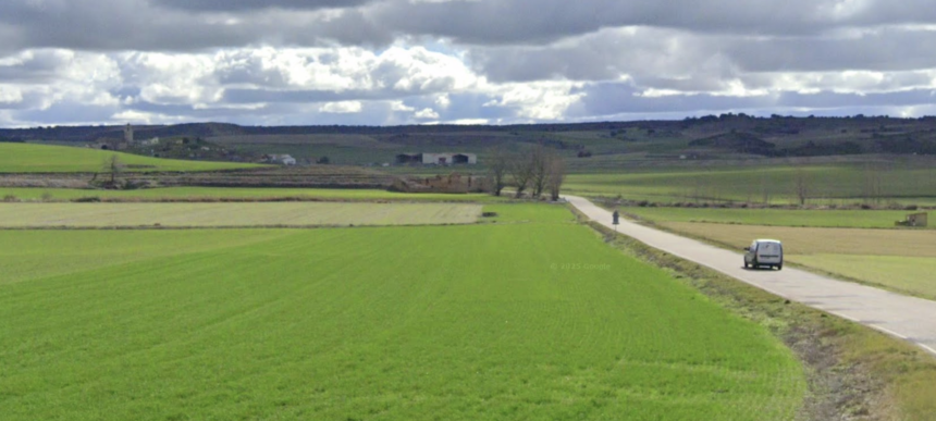 Furgoneta circulando por una carretera provincial entre campos de cultivos con un conjunto de casa al fondo.