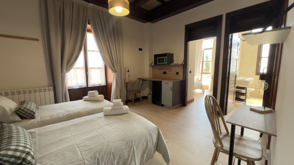 Habitación con dos camas individuales con toallas sobre ellas, un pequeño office con microondas, un escritorio y dos puertas que dan acceso al baño y a otr habitación.