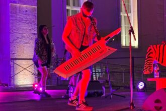 Un joven con pantalón u gafas de sol y un piano-guitarra toca sobre un escenario