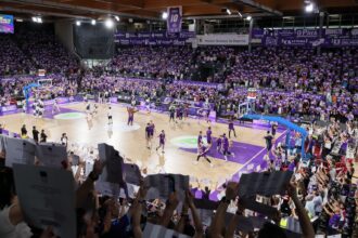 cientos de aficionados sostienen pancartas moradas mientras en la cancha los jugadores de baloncesto calientan