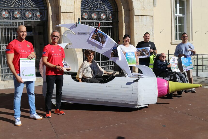 Varias personas posan delante del Ayuntamiento con distintos carteles de las actividades que se van a organizar. La concejala está sentada en un vehículo de creación casera o autoloco.
