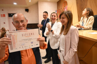 Un hombre muestra un diploma ante tres personas que sonrien.